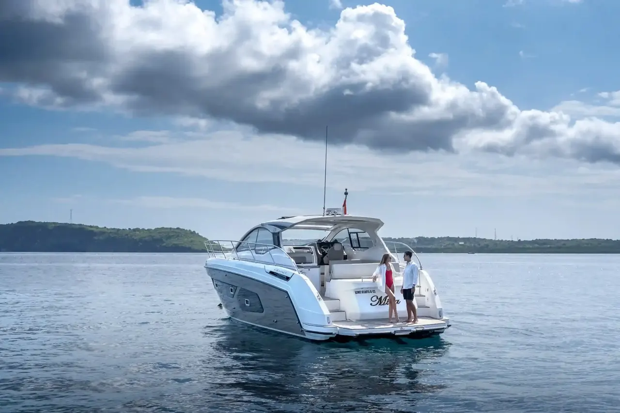 Two guests at the stern of a white motor yacht anchored off a green headland