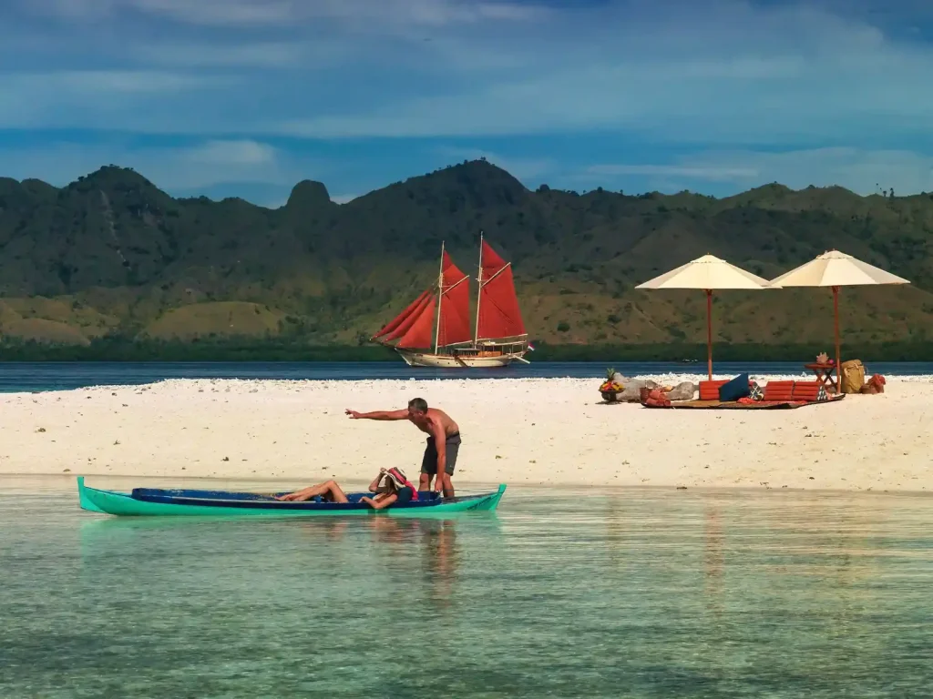 A red-sailed phinisi off a white beach in Komodo National Park